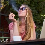 Woman smoking outside with laptop and coffee.
