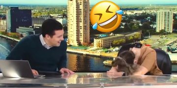 News anchors laughing during broadcast with city backdrop.
