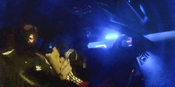 Masked person eating sandwich in dark car, blue light.