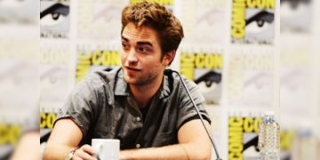 Person at Comic-Con panel holding mug, speaking.