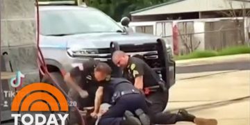 Police restrain person on ground near vehicle.