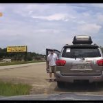 Man exits silver SUV near roadside sign.