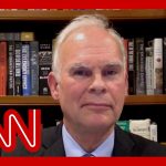 Man in suit with bookshelf background, CNN logo overlay.