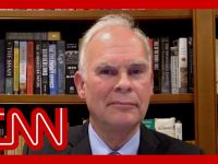 Man in suit with bookshelf background, CNN logo overlay.