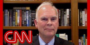 Man in suit with bookshelf background, CNN logo overlay.